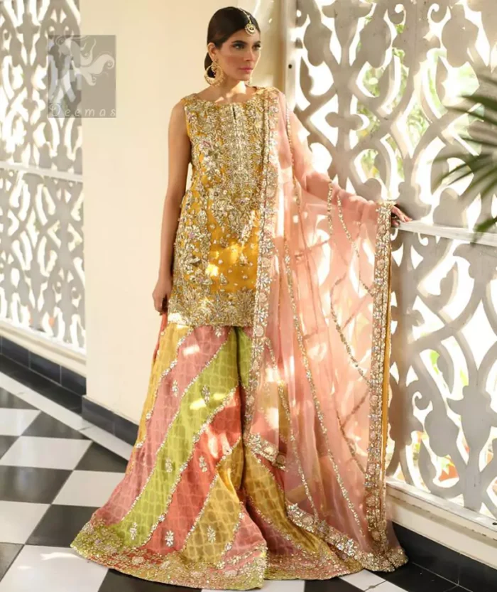 Amber shirt adorned with mainly light golden embellishment on neckline, hemline and side slits. Pink peach dupatta adorned with embellished border and stripes. Multiple color sharara adorned with border on hemline, stripes and motifs.
