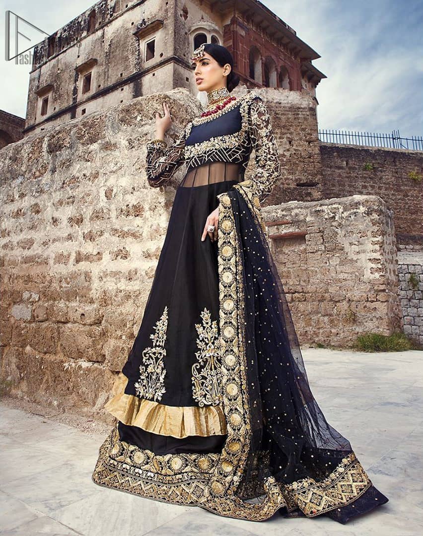 Delicately crafted and personifying chic elegance with an element of grandiose. This regal black outfit is an immensely captivating traditional piece, enhancing the art of classical heritage showcasing the craftsmanship of golden kora, dabka, tilla detailed with sequins and pearls; artistically embellished to give a beautiful rhythm to the outfit. Furthermore the anarkali frock is enhanced with fascinating embellishment on neckline and frilled hemline. It comes with an exquisite black lehenga with thick embroidered bottom to give it a regal look. It comes with a black net dupatta with sequins sprayed all over finished with thick embellishment all around the edges.