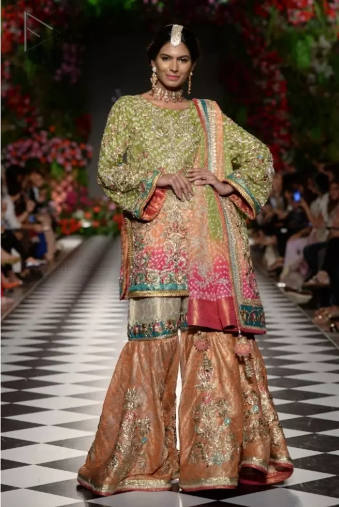 Brighten up your look with this beautiful mehndi dress comes with a beautiful parrot green shirt. This shirt is adorned with light gold kora, dabka, tilla and sequins work, furthermore the hemline is enhanced with applique embellishment. Paired it up with traditional jamawar gharara ornamented with zardozi work and finished with lace embellished border. It is coordinated with parrot green dupatta dyed from pallu in two colors and adorned with applique embellished border.