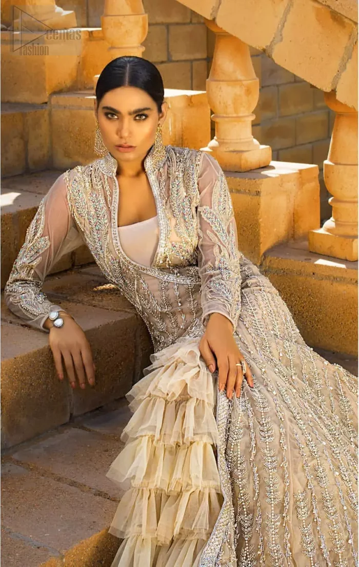 A fusion of traditional and latest fashion making the outfit trendy and unique. This angrakha style floor length maxi is heavily hand crafted with floral embroidery on the bodice, floral booties and vertical lines all over the dress. Back is also enhanced with vertically designed floral booties and swarovski crystals lines all over. The embroidery is done in the shades of silver and dull golden zardozi work. Style it up with frilled lehenga to give it a regal look. This outfit is comprises with fawn net lehenga sprinkled with sequins spray all over.