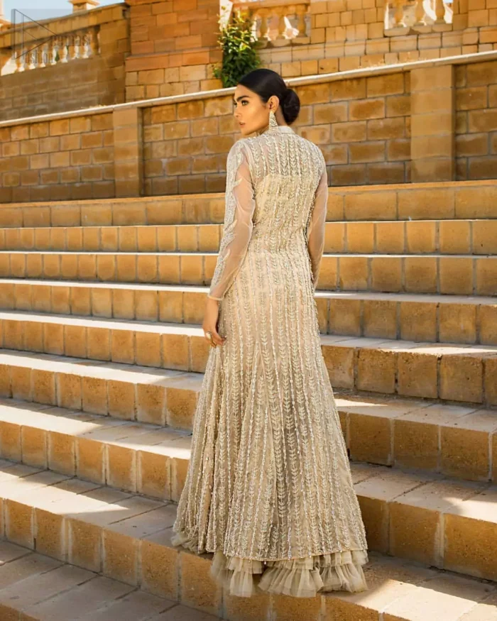 A fusion of traditional and latest fashion making the outfit trendy and unique. This angrakha style floor length maxi is heavily hand crafted with floral embroidery on the bodice, floral booties and vertical lines all over the dress. Back is also enhanced with vertically designed floral booties and swarovski crystals lines all over. The embroidery is done in the shades of silver and dull golden zardozi work. Style it up with frilled lehenga to give it a regal look. This outfit is comprises with fawn net lehenga sprinkled with sequins spray all over.