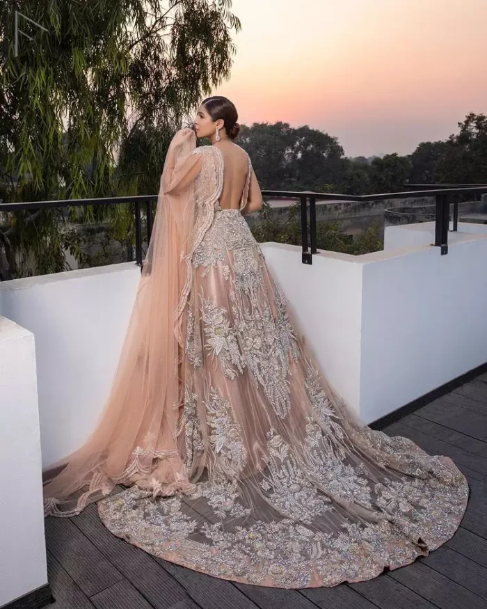 Some brides definitely shine bright by stepping out of the classic box and jumping into pastel hues for their big day. The front open back train dreamy maxi is adorned with an embellished bodice and floral booties all over the body giving a perfect ending to this outfit. The embroidery is done in the shade of silver and gold. Furthermore, the back train is emphasized with a large central motif and thick embellished borders. Coordinated with matching net lehenga and tea rose dupatta having a four-sided scalloped border.