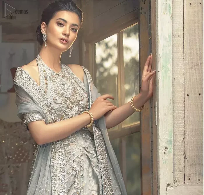 Nikah Wear - Light Grey Halter Neck Gown Lehenga. The floral and the flowery climber networks on fully embellished Halter neck and blouse has exquisitely exaggerated in the beauty of the costume making it very unique. The bottom lehenga with Fixed Waist Belt With Side Zip Closure is composed of Katan Banarsi Jamawar fully embellished with flowery climbers with Light Gray colour tone; being exquisitely beautiful in perfecting the complete outfit having a unique fabric and double flare lehenga that is in stellar combo with the blouse with finished edges and Dupatta sideways carried beautifully by the bride with organza fabric and Heavy beautiful golden and Grayish lace on all four bordered finished sides and dabka work on the net inside of the gorgeously made dupatta, whole this costume has its own unifying feature such as 100 % imported high quality overall highly embellished fabric making it the best choice to have by you on your big day.
