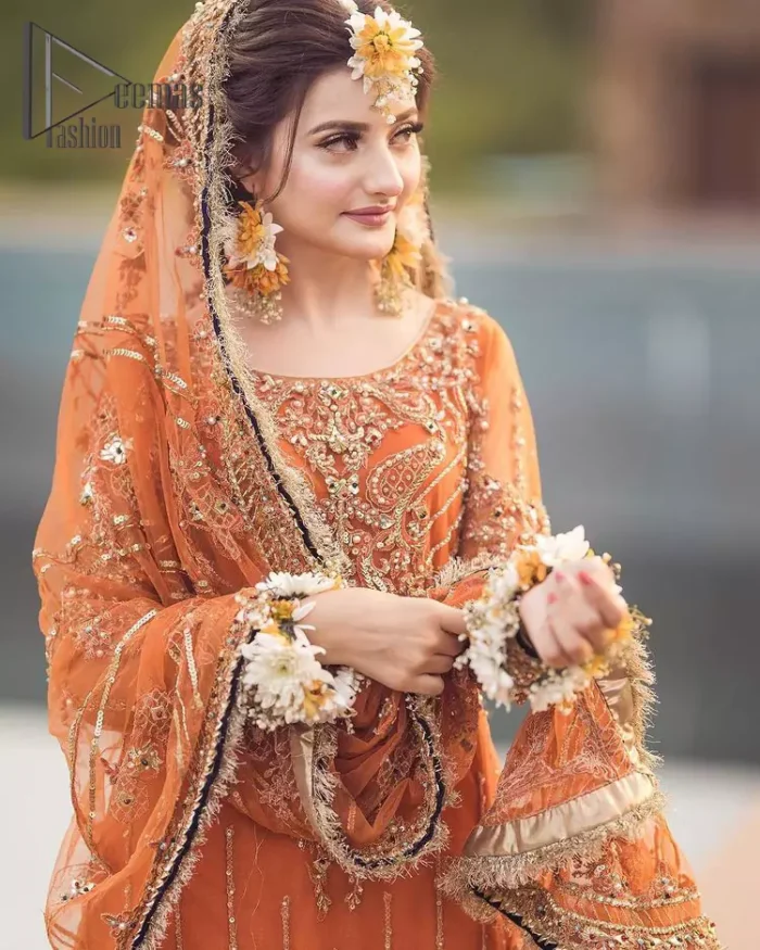 Time to treat yourself with an orange article of Deemas Fashion. This orange heartiest mealtime article will twirl your heart.  Prettify your Mehndi look with this orange short shirt that is embellished with tilla, kora and dabka work. Its rounded neckline with full sleeves gives you a genuine feeling of a bride. It is synchronized with flayer orange sharara that has an embellished border and spray of tiny floral motifs that gives a super aesthetic look to this dress. Finish this look with a net dupatta that has four sides embellished border and Kiran work.
