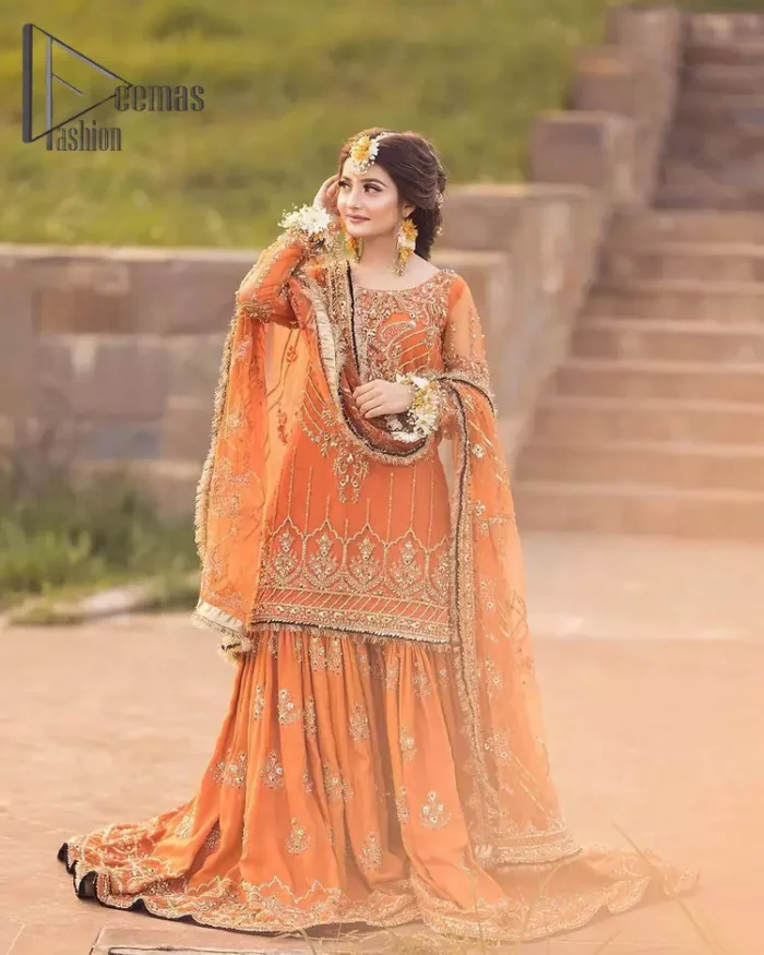 Time to treat yourself with orange article of Demaas Fashion. This orange heartiest mealtime article will twirl your heart.  Prettify your Mehndi look with this orange short shirt that is embellished with tilla, kora and dabka work. Its rounded neckline with full sleeves gives you a genuine feeling of bride. It is synchronize with flayer orange sharara that have embellished border and spray of tiny floral motifs that gives a super aesthetic look to this dress. Finish this look with net dupatta that have four side embellished border and kiran work.