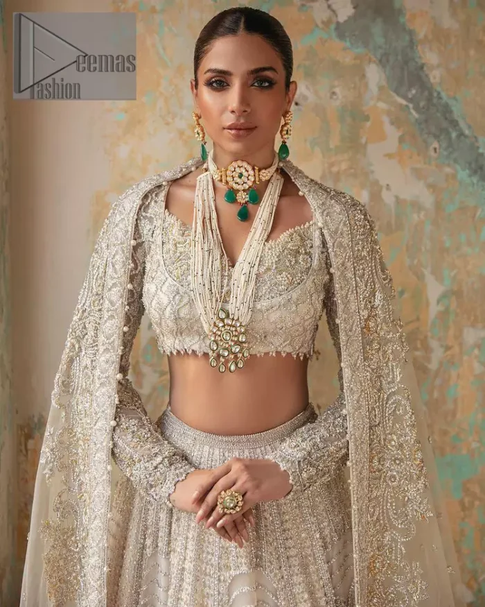With a touch of Divine. A stunning ivory blouse is laboriously frosted with meticulously detailed hand embellishment with kora, dabka, tilla and pearls all over. The sweetheart neckline of the blouse and full sleeves together gives a so romantic look to any bride. In addition to this, it is systemized with the same colour frilled lehenga which is laden with the same colour embroidery that gives a magical stream of tilla, dabka and pearls. Finish this ivory article with the same colour dupatta that is heavily embellished with four-sided handsome borders and sequins sprayed all over to enhance the feeling of joy. 