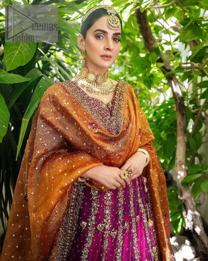 The outfit defines the true essence of allure with its spellbinding magenta hue and intricate details. The blouse is decorated with golden embroidery based on the neckline and sleeves. The sleeves are further enhanced with the embellished handwork. It is Beautifully organized with a magenta lehenga that is again heavily embellished with golden embroidery that is just adding poise to the whole look. Paired along with an orange dupatta decorated with subtly adorned motifs and embellished border that is completing this Mehandi looks wondrously.