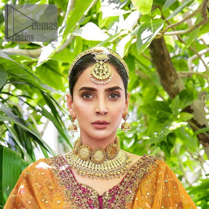 The outfit defines the true essence of allure with its spellbinding magenta hue and intricate details. The blouse is decorated with golden embroidery based on the neckline and sleeves. The sleeves are further enhanced with the embellished handwork. It is Beautifully organized with a magenta lehenga that is again heavily embellished with golden embroidery that is just adding poise to the whole look. Paired along with an orange dupatta decorated with subtly adorned motifs and embellished border that is completing this Mehandi looks wondrously.