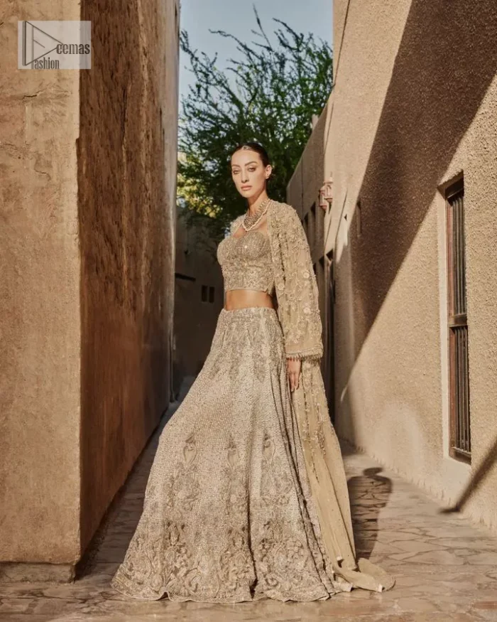 This modern piece is all about extraordinary details and aesthetic vibes! A beige organza blouse is heavily embellished with dabka, naqshi, tilla, Resham, dabka, sequin and Kundan. This gown blouse dress has Mughal-inspired motifs, geometrical patterns and floral motifs. The blouse has a sweetheart neckline or a more upscale appearance and feel. Further, the gown has full sleeves adorned with floral patterns. It is supported by a flared lehenga with an embellished border for additional comfort.