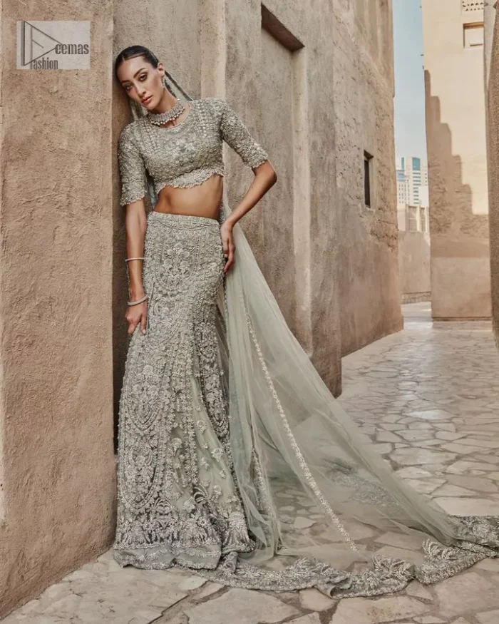 This fusion of floral embroidery is total fashion goodness for a walima outfit! A stunning teal grey organza choli embellished with intricate detailing finished with a scalloped border. The beautiful blouse is decorated with silver embellishment which is further prominent with tilla,daka, kora, Kundan and the real magic of Zardozi.  It is paired with a heavily embellished back-train lehenga with botanical motifs and geometric patterns. Its dupatta is made from a net. The dupatta is a wide embellished border that added divine charm to the entire ensemble.