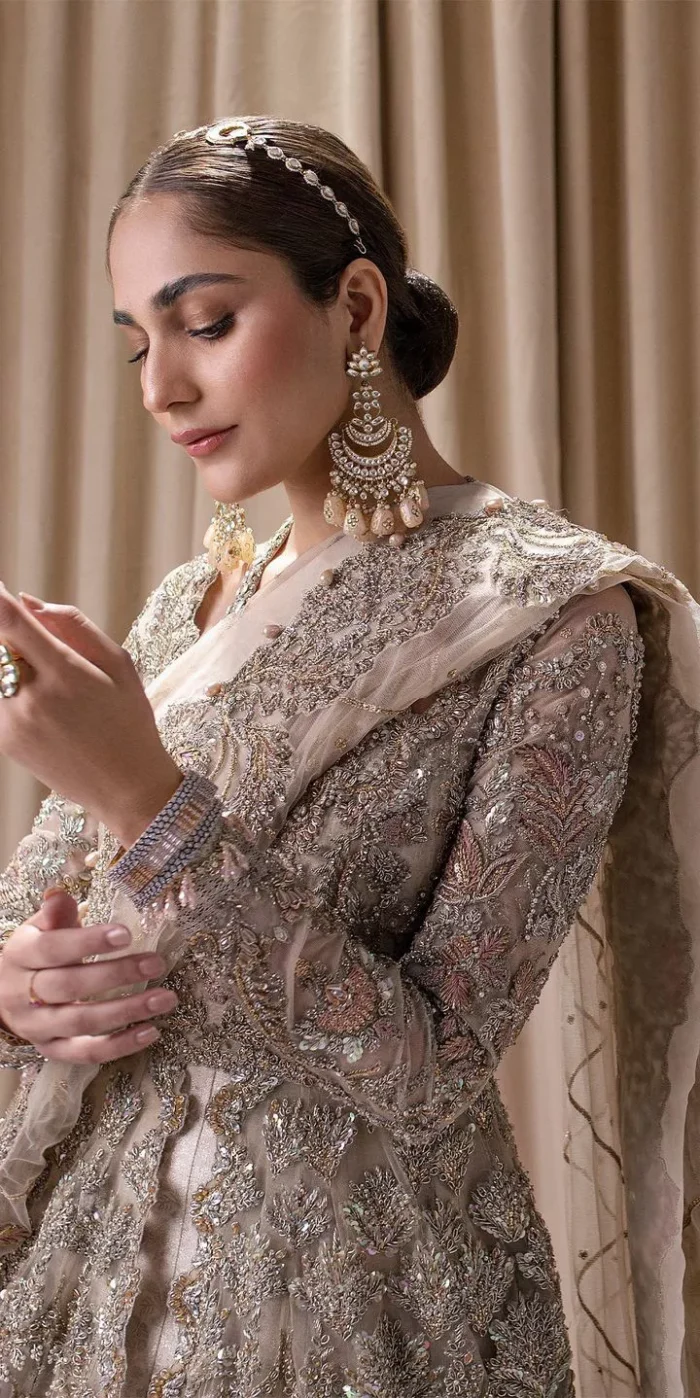 Make a statement with a light fawn article on your nikah day! DeemasFashion presents the light fawn heavy maxi which is embellished with silver embroidery using dabka, tilla, kora, Zardozi, Swarovski, Mukesh, sequin and Kundan. The strap neckline adds a little romanticness to the dress. Further, the floral patterns on full sleeves make this masterpiece modern and unique. It is paired with a  lehenga, embellished with a delicate border. It is styled with a dupatta; its surface is sprayed with sequin and the dupatta is enveloped by a heavily embellished floral border.