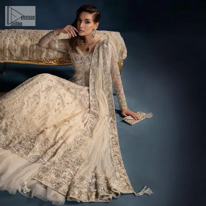 A sophisticated splash of off-white as your nikah outfit! With its round neckline and full sleeves, this outfit has a fine fusion look. Utilizing attractive silver embellishment work to enhance its intricate beauty. Tilla, dabka, kora, Kundan and the details of Zardozi beautifully embellish the elegant frock. It is paired with a flared lehenga to balance the overall look of this charming article. Finish this soft and bold look with a heavily bordered four-sided dupatta with a spray of sequins all over to make your Big day super aesthetic.